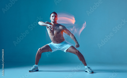 Zumba movement by young healthy strong white man with bare body. Male dancer wearing white shorts and sneakers, standing on blue background.