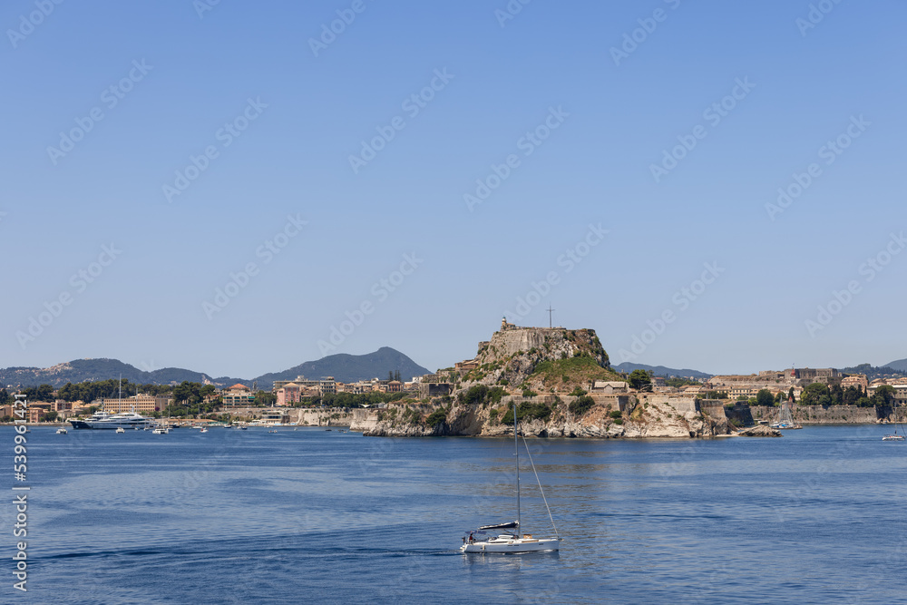 Lonely small white yacht with folded sails against the backdrop of huge Old Fortress dominating all region, Corfu island, Greece, sunny morning