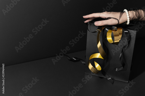 Woman with chiffon glove holds shopping bag with golden streamers in dark studio closeup. Black Friday holiday discounts for customers (ID: 539964461)