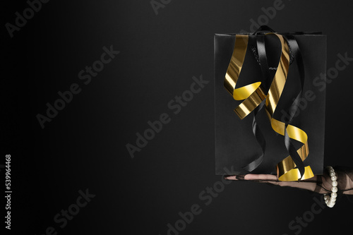 Woman shows shopping bag with shiny golden streamers in dark studio closeup. Black Friday holiday sale. Fashion industry advertisement (ID: 539964468)