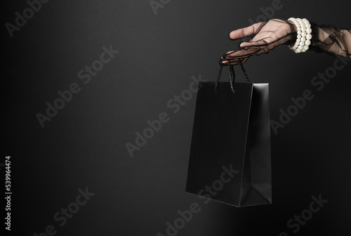 Blank shopping bag hangs on woman finger in dark studio closeup. Presentation of luxury goods during Black Friday sales. Discount season (ID: 539964474)