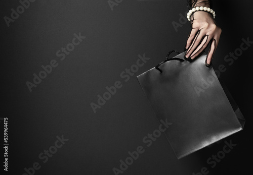 Stylish shopping bag held by woman hand in dark studio closeup. Special offers for Black Friday holiday. Great sales and discount system (ID: 539964475)