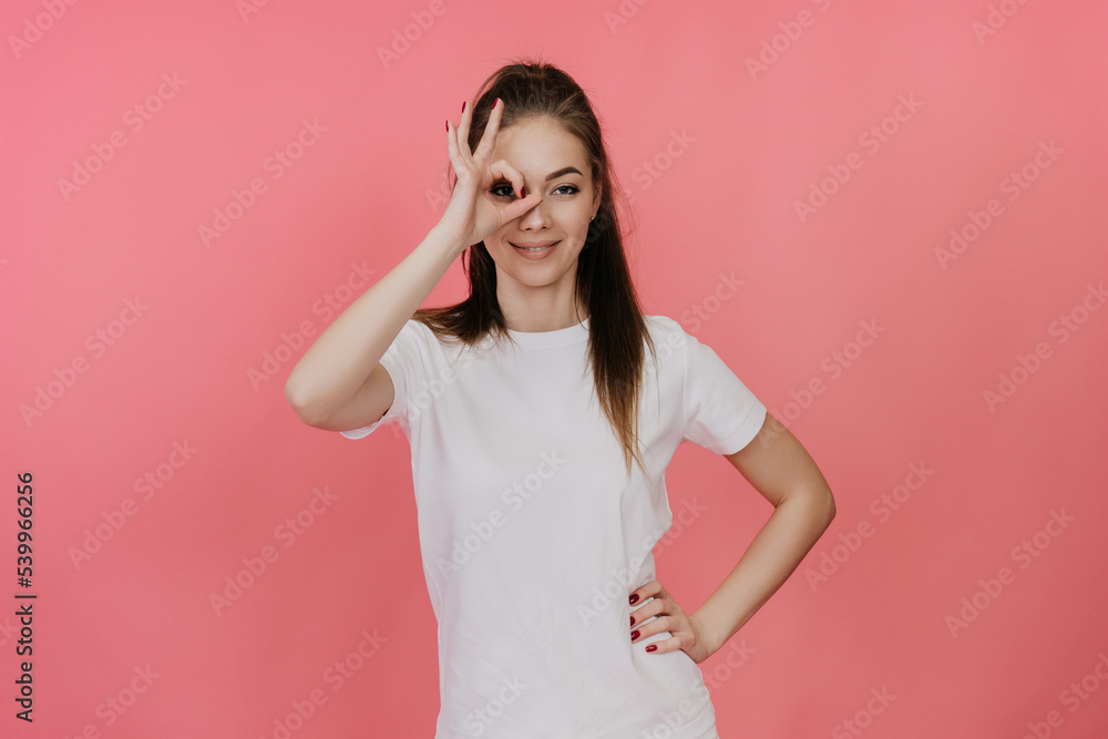Playful young blonde in a white T-shirt looking through a hand-shaped gesture against pink background. Cheerful student girl making ok sign, satisfied by self development and education. Youth concept. Playful young blonde in a white T-shirt looking through a hand-shaped gesture against pink background. Cheerful student girl making ok sign, satisfied by self development and education. Youth concept.