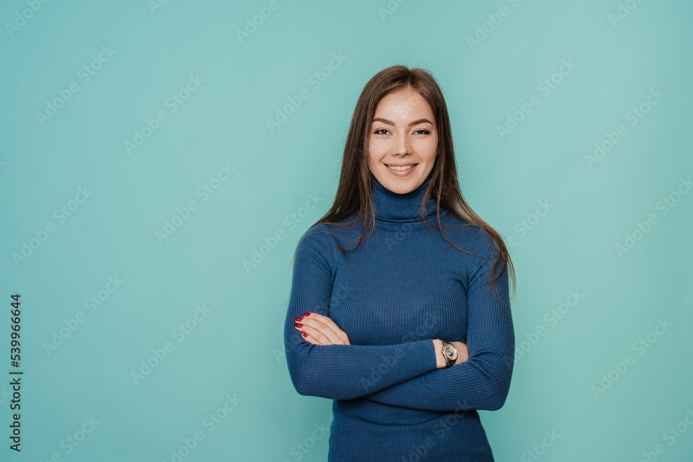 Beautiful blonde young woman with loose hair in blue sweater folding hands on chest looking at camera toothy smiling over turquoise studio backdrop. Pretty Italian confident female. Mockup. Beautiful blonde young woman with loose hair in blue sweater folding hands on chest looking at camera toothy smiling over turquoise studio backdrop. Pretty Italian confident female. Mockup.