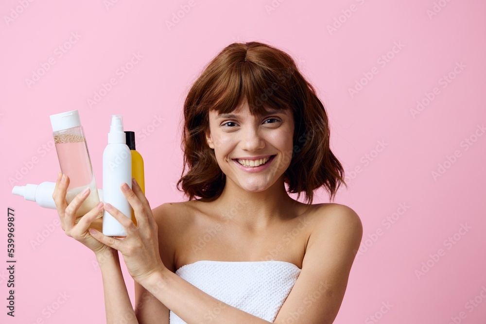 a happy, young woman stands on a pink background, wrapped in a white towel and holding a set of jars with facial care cosmetics in her hands, looking widely at the camera a happy, young woman stands on a pink background, wrapped in a white towel and holding a set of jars with facial care cosmetics in her hands, looking widely at the camera