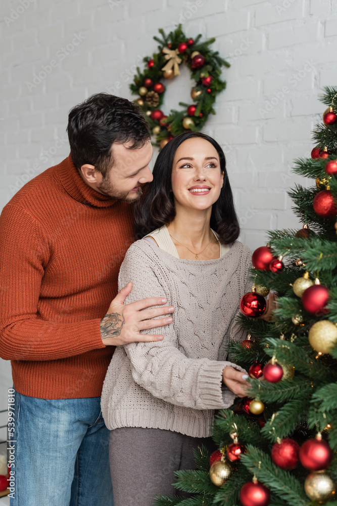 man hugging cheerful brunette wife decorating christmas tree at home man hugging cheerful brunette wife decorating christmas tree at home