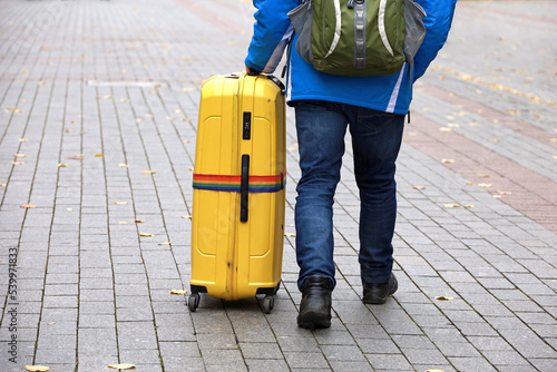 Man in jeans walking with backpack and big yellow suitcase on wheels. Male legs and luggage on autumn street, travel concept