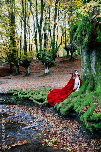 A woman in a white dress and a long red cape is hiding behind a tree in the middle of the forest next to a stream.