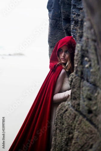A woman in a white dress with a long red cape and hood hides from someone among the rocks.