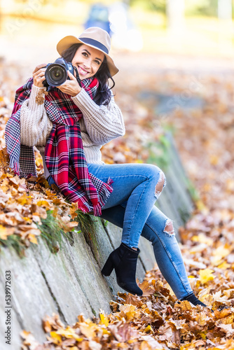 Smiling good-looking female photographer in a hat and stylish autumn clothes takes pictures outside on a sunny fall day