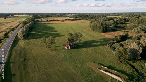 Aerial drone flyover baltic country side of Estonia, Europe with a orthodox or catholic lost place church ruin without roof on a green meadow next to country road with cars on a hot summer day 2022