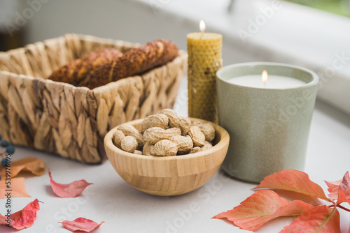 Still life candle flame, peanut, fallen leaves, wicker basket, bagel on a table, home decor in a cozy house. Autumn weekend concept 
