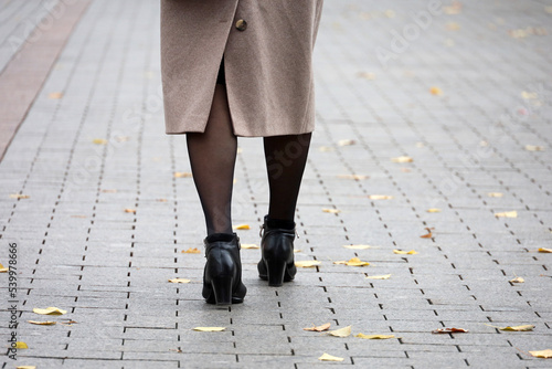 Female legs in black stockings and shoes on high heels on paved sidewalk. Woman in coat walking down the street covered fallen leaves, fashion in autumn city