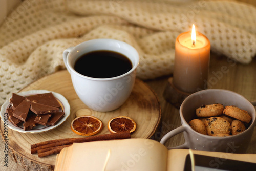 Bowl of cookies, cup of tea or coffee, chocolate, spices, knitted blanket, books, glasses and candle on the table. Cozy hygge atmosphere at home. Selective focus.
