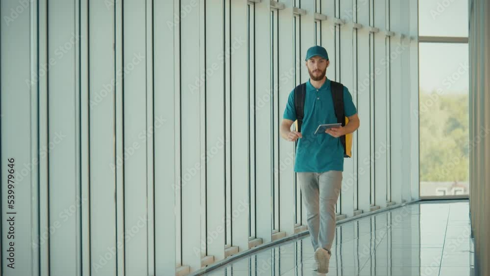 Food Delivery Person in Uniform and With a Thermal Backpack Walking in Modern Office Building and Using Tablet to Check Order. Courier On the Way to Deliver Order to a Client. Food Delivery Person in Uniform and With a Thermal Backpack Walking in Modern Office Building and Using Tablet to Check Order. Courier On the Way to Deliver Order to a Client.