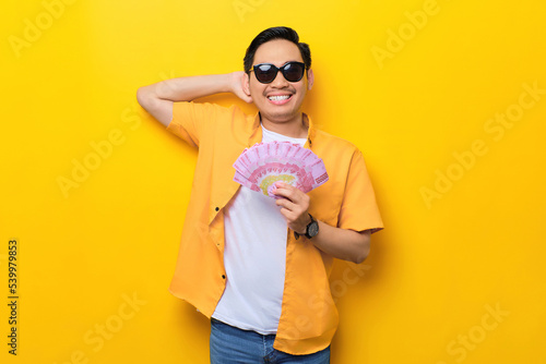Cheerful young handsome Asian man wearing sunglasses holding fan of money banknotes isolated on yellow background