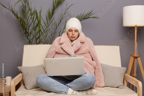 Photography Horizontal shot of sad Caucasian woman posing in cold room sitting on the sofa wearing warm hat, mittens and cap, holding laptop on knees, looking at display with pout lips