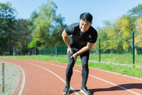 young asian runner athlete with muscle pain. Man massaging Stretching, trauma injury while jogging at the stadium outdoors. Fitness male sprain severe pain stretch pull. Leg muscle cramp calf sport