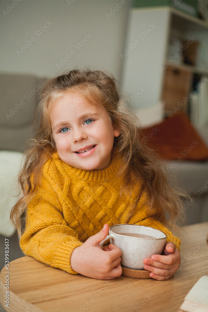 Cute little blue-eyed curly girl 4 years old drinking tea in a cozy house. Autumn. Hot drink. Cute little blue-eyed curly girl 4 years old drinking tea in a cozy house. Autumn. Hot drink.