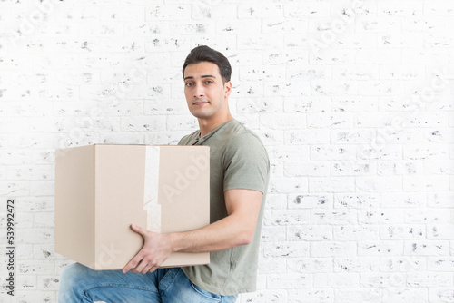 Handsome man carrying huge and heavy box in front of white brick wall background 