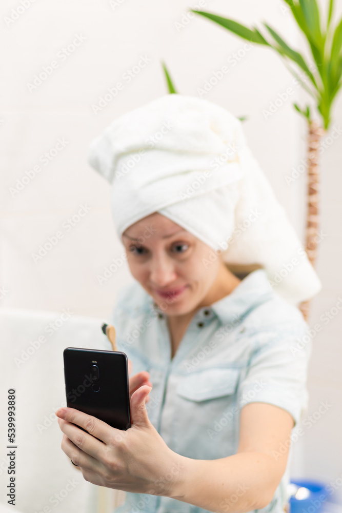A young pretty woman with a towel on her head in a bright bathroom with a toothbrush and a smartphone in her hand brushes her teeth and communicates in social networks. Selective focus. Close-up A young pretty woman with a towel on her head in a bright bathroom with a toothbrush and a smartphone in her hand brushes her teeth and communicates in social networks. Selective focus. Close-up