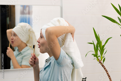 A young pretty woman in a blue bathrobe with a towel on her head in a bright bathroom with a toothbrush in her hand sings songs. Reflection in the mirror with a green houseplant.