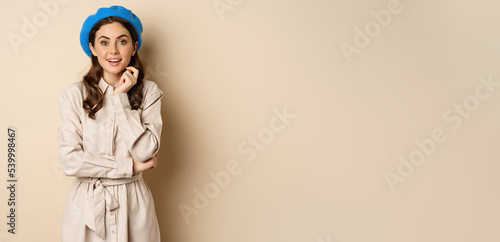 Image of stylsh beautiful woman looking surprised, shocked reaction at camera, posing in trench coat against beige background photo