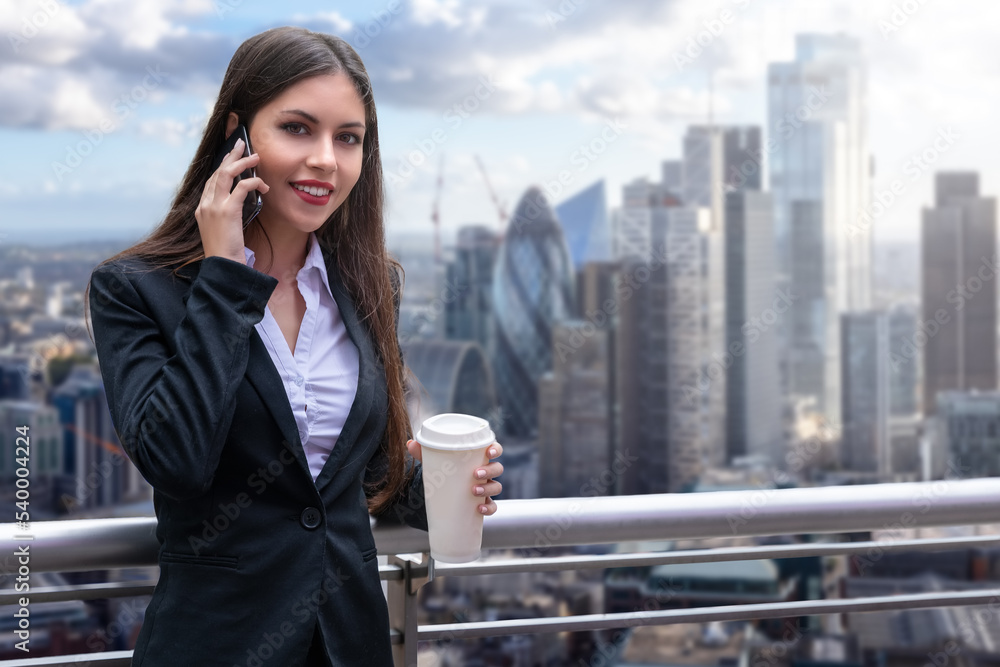 A attractive business woman in a corporate outfit holding a coffee cup talks on her mobile telephone in front of the city skyline of London, England A attractive business woman in a corporate outfit holding a coffee cup talks on her mobile telephone in front of the city skyline of London, England