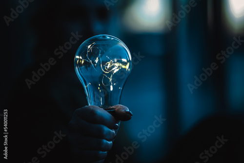 young worried woman holding bright shining lightbulb in her hands feeling unsave while in fear thinking about a future blackout with no electricity or power outage with blue cold background