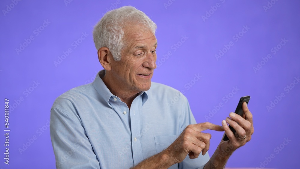 Happy fun elderly man 70s in blue shirt gets video call using mobile cell phone talk wave with hand isolated on plain light purple background studio portrait Happy fun elderly man 70s in blue shirt gets video call using mobile cell phone talk wave with hand isolated on plain light purple background studio portrait