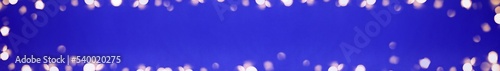 Banner of Christmas bright gold colors bokeh different sizes around the frame on vibrant blue blurred background. Holiday template and shiny greeting concept with copy space.