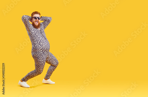 Crazy funny fat man with big belly in pajama suit with leopard print is dancing and fooling around. Cheerful chubby bearded freak man in sunglasses laughing loudly on orange background. Full length.