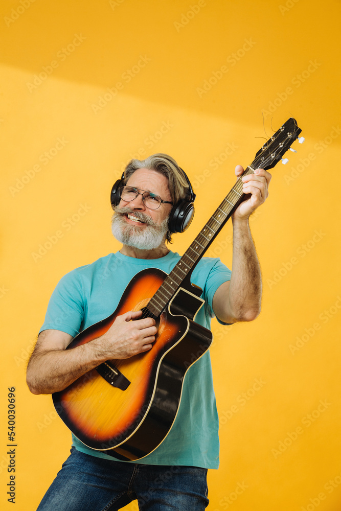 Senior man in glasses and headphones playing the guitar on a yellow background. Studio shooting.