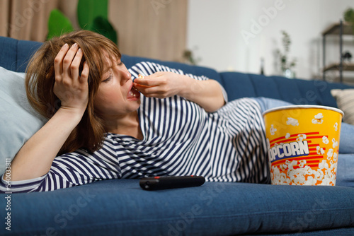 Upset lonely woman liyng on sofa eating popcorn and watching boring tv programmes, television consumerism