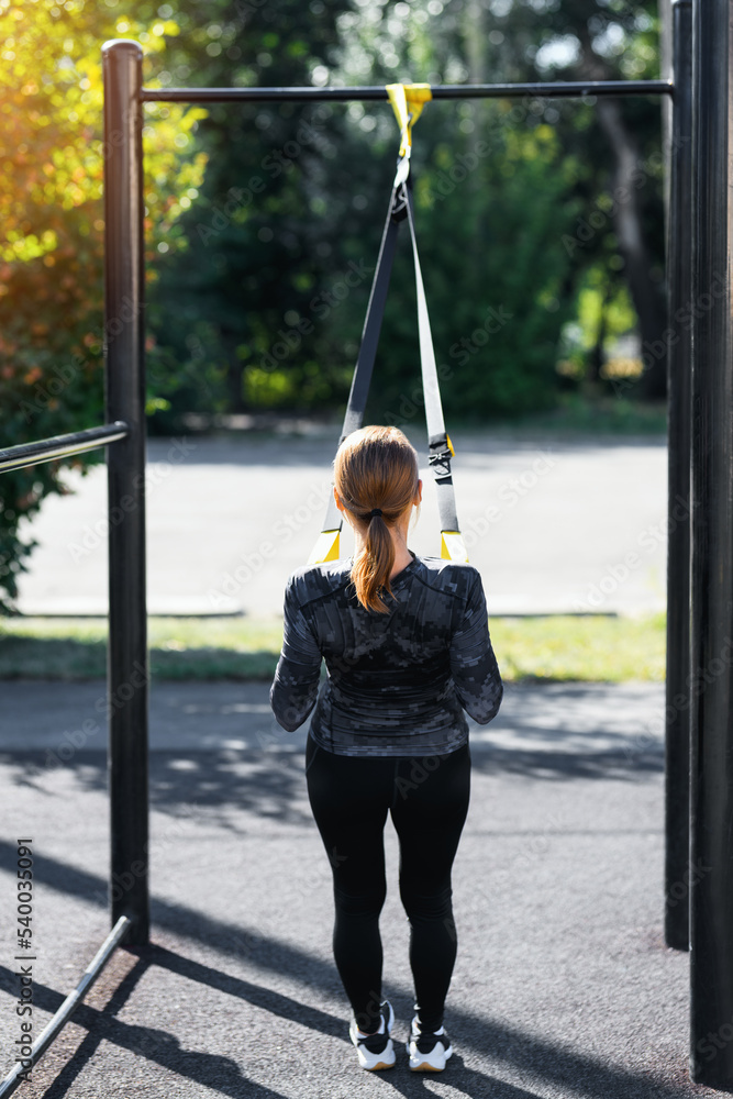 Women and sport. Girl in sportswear with sports equipment TRX exercising outdoors at the gym, view from the back. Middle aged sportswoman Women and sport. Girl in sportswear with sports equipment TRX exercising outdoors at the gym, view from the back. Middle aged sportswoman