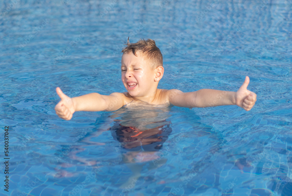 Little blond boy in the outdoor pool is showing thumbs up