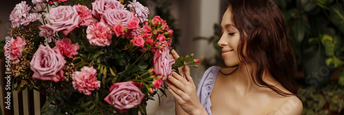 Banner portrait of a beautiful young brunette woman in a purple dress. She is holding a vintage vase with a large spring bouquet of pink natural flowers.