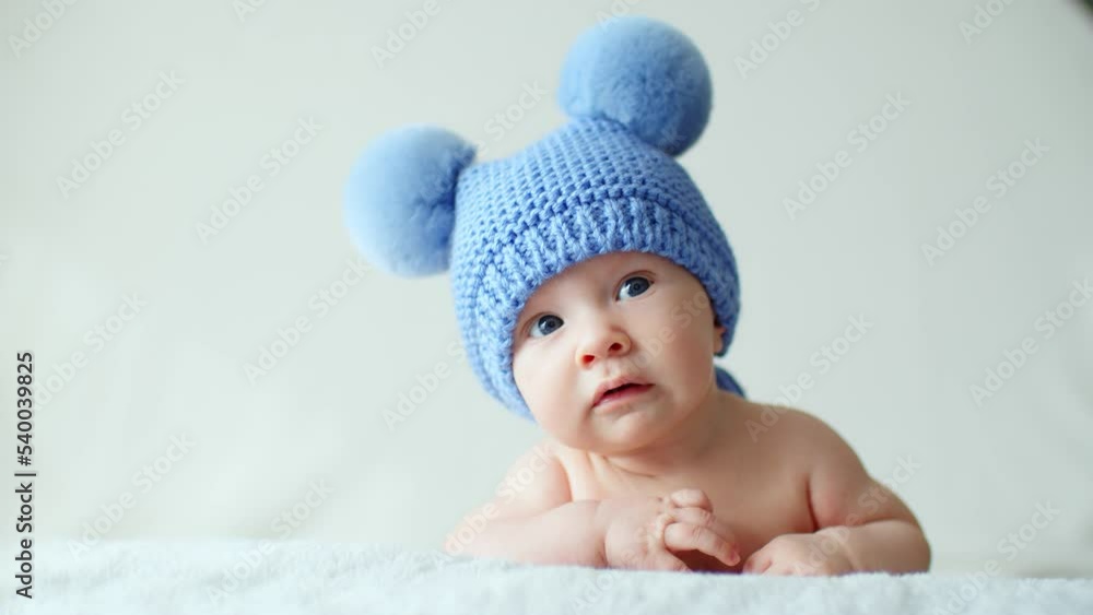 Calm two-month-old infant is lying on a blanket. Beautiful, cute little child with blue eyes looks into camera. Sweet newborn baby in a blue hat with pompoms looks into the camera. SLOW MOTION Calm two-month-old infant is lying on a blanket. Beautiful, cute little child with blue eyes looks into camera. Sweet newborn baby in a blue hat with pompoms looks into the camera. SLOW MOTION