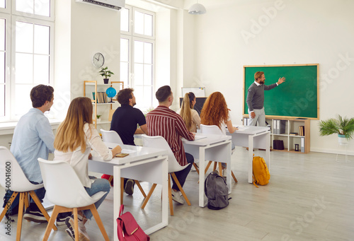 Group of students enthusiastically and attentively listens to professor pointing green chalkboard. Modern, comfortable, ergonomic school space. Seminar, colloquium at university.