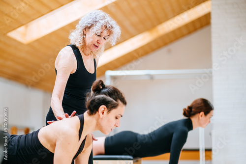 Pilates lesson on chair, side view of blond elderly personal trainer coaching two young women indoor at studio.