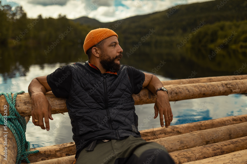An african american man with an orange beanie sitting on a wooden raft on a wide river watching the water.