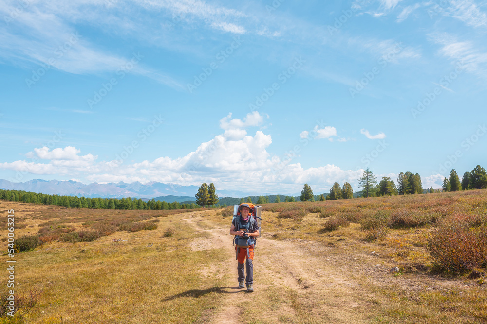 Alone traveler with large backpack walks along hiking trail on sunlit high mountain plateau under white clouds in blue sky. Backpacker with photo camera in autumn mountain trekking in good weather.