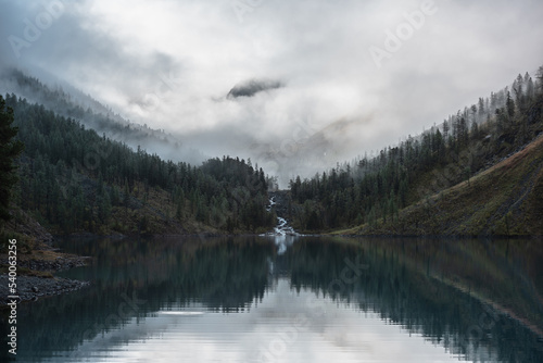 Mountain creek flows from forest hills into glacial lake. Tranquil scenery with rocks in clearance of mysterious fog. Small river and coniferous trees reflected in calm alpine lake in early morning.