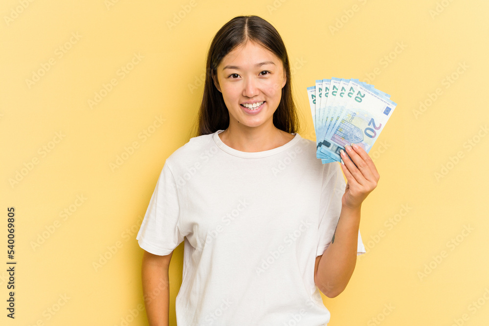 Young asian woman holding a banknotes isolated on yellow background happy, smiling and cheerful. Young asian woman holding a banknotes isolated on yellow background happy, smiling and cheerful.