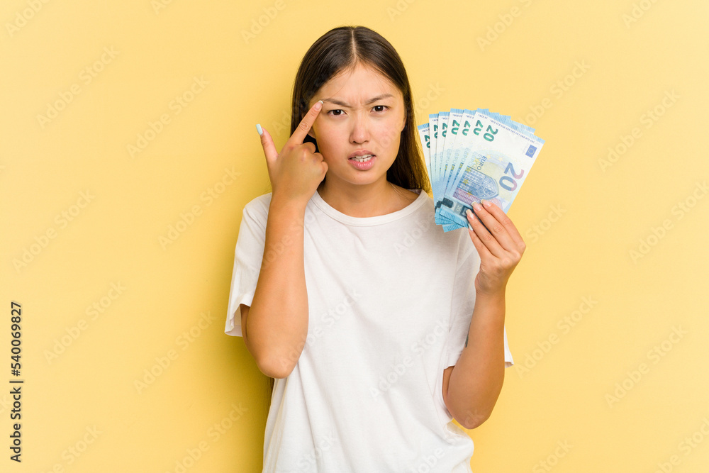 Young asian woman holding a banknotes isolated on yellow background showing a disappointment gesture with forefinger. Young asian woman holding a banknotes isolated on yellow background showing a disappointment gesture with forefinger.