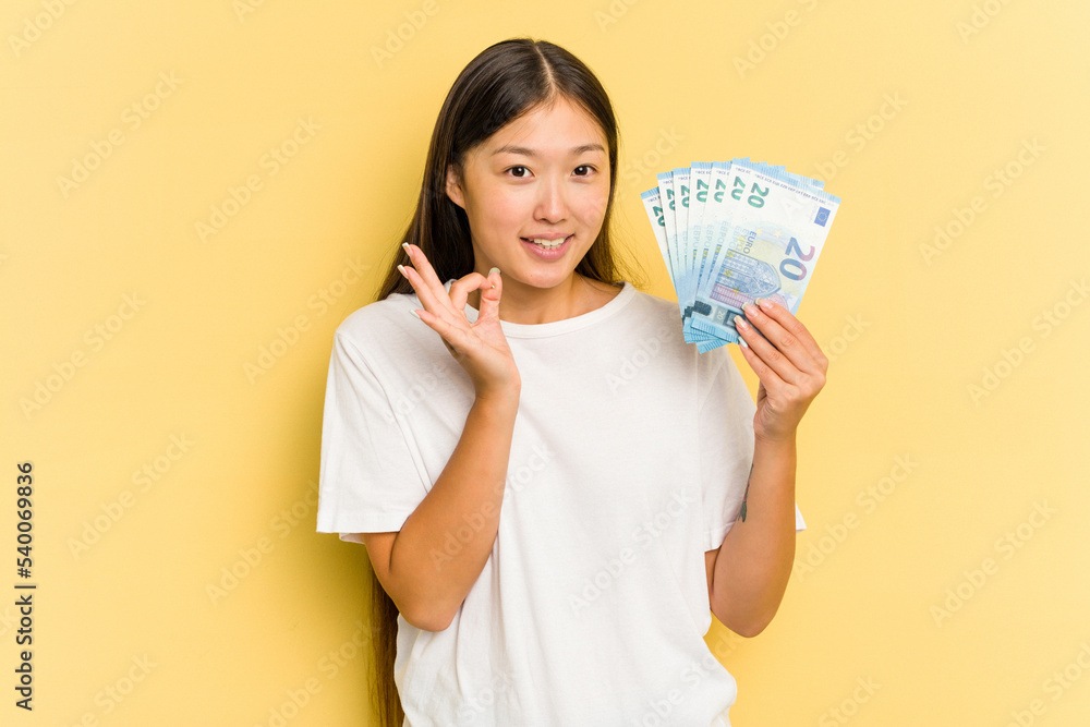 Young asian woman holding a banknotes isolated on yellow background cheerful and confident showing ok gesture. Young asian woman holding a banknotes isolated on yellow background cheerful and confident showing ok gesture.