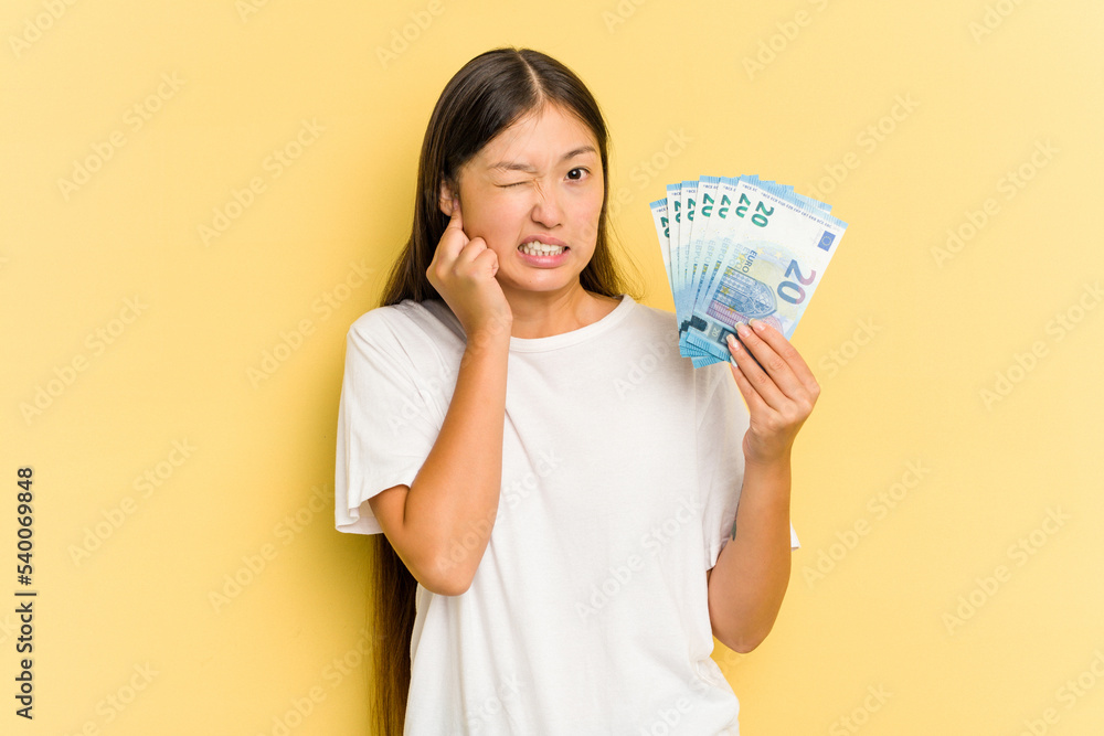 Young asian woman holding a banknotes isolated on yellow background covering ears with hands. Young asian woman holding a banknotes isolated on yellow background covering ears with hands.