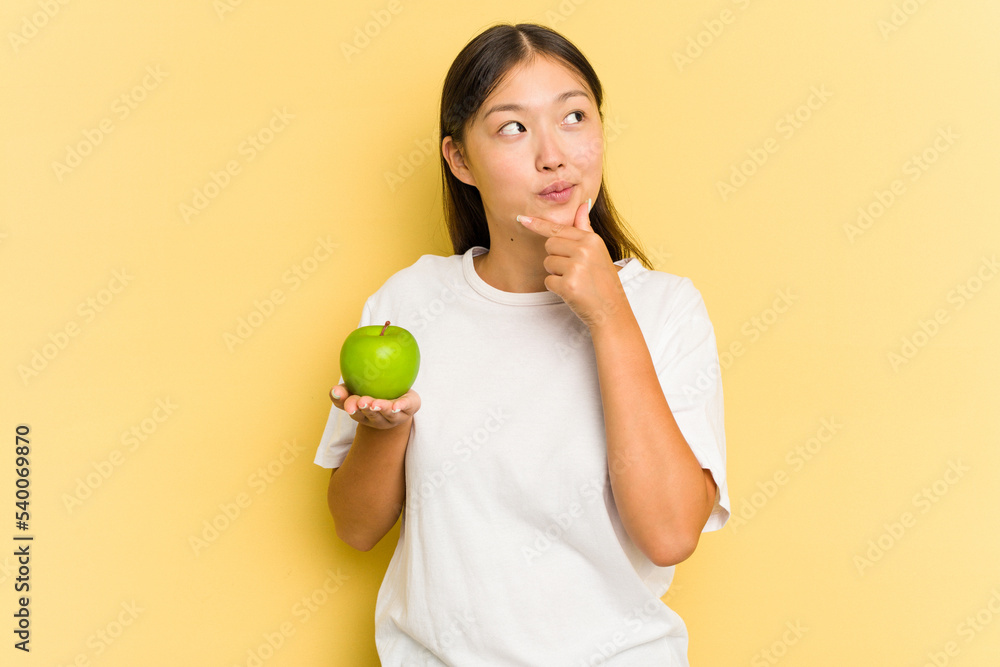 Young Asian woman eating an apple isolated on yellow background looking sideways with doubtful and skeptical expression. Young Asian woman eating an apple isolated on yellow background looking sideways with doubtful and skeptical expression.