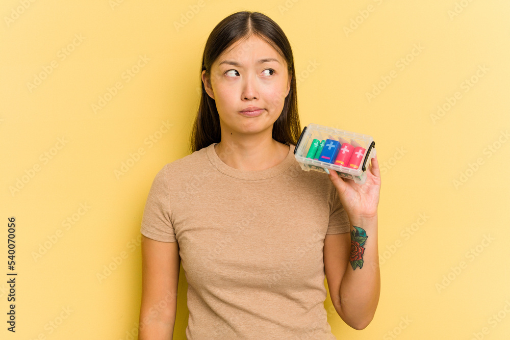 Young Asian woman holding batteries to recycle them isolated on yellow background confused, feels doubtful and unsure.