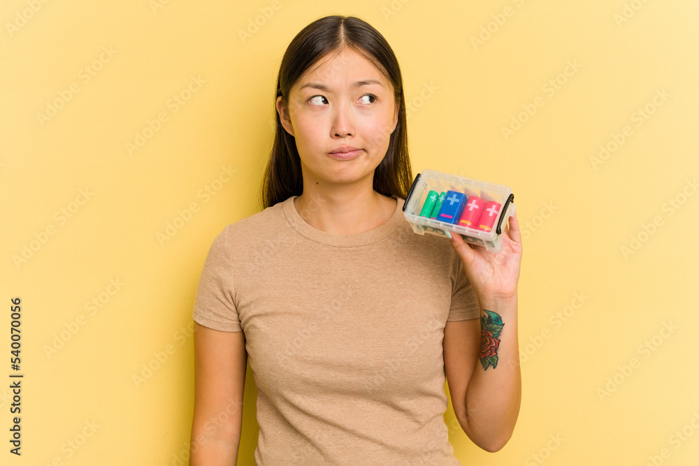 Young Asian woman holding batteries to recycle them isolated on yellow background confused, feels doubtful and unsure. Young Asian woman holding batteries to recycle them isolated on yellow background confused, feels doubtful and unsure.